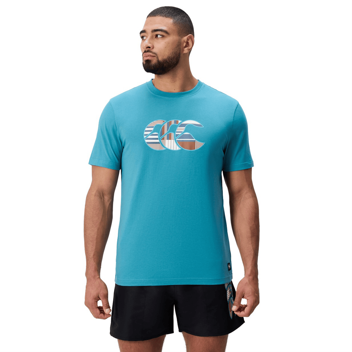 A man stands facing forward with arms at his sides, wearing a teal Canterbury Uglies Logo T-shirt in a soft cotton blend featuring a large logo on the chest, paired with black athletic shorts.
