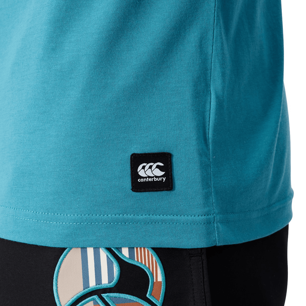 Close-up of a person in a blue Canterbury Uglies Logo T-shirt made from a soft cotton blend, featuring the bold Canterbury logo. They're paired with black shorts accented by colorful designs near the hem.