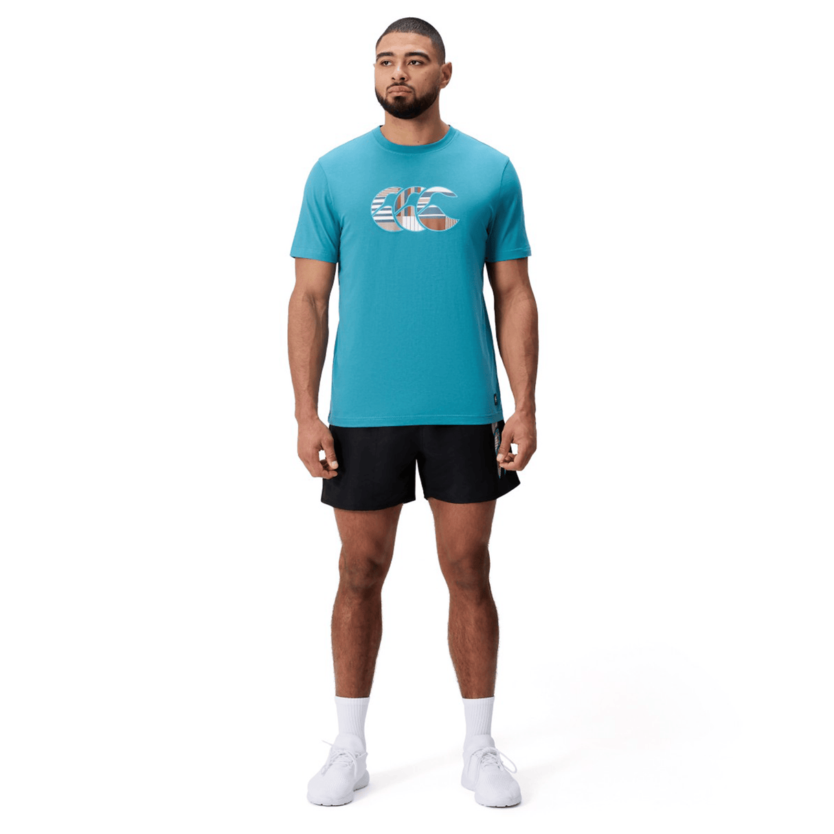 A man faces forward wearing a Canterbury Uglies Logo T-shirt in teal cotton blend, paired with black shorts and white athletic shoes, all photographed against a plain white background.