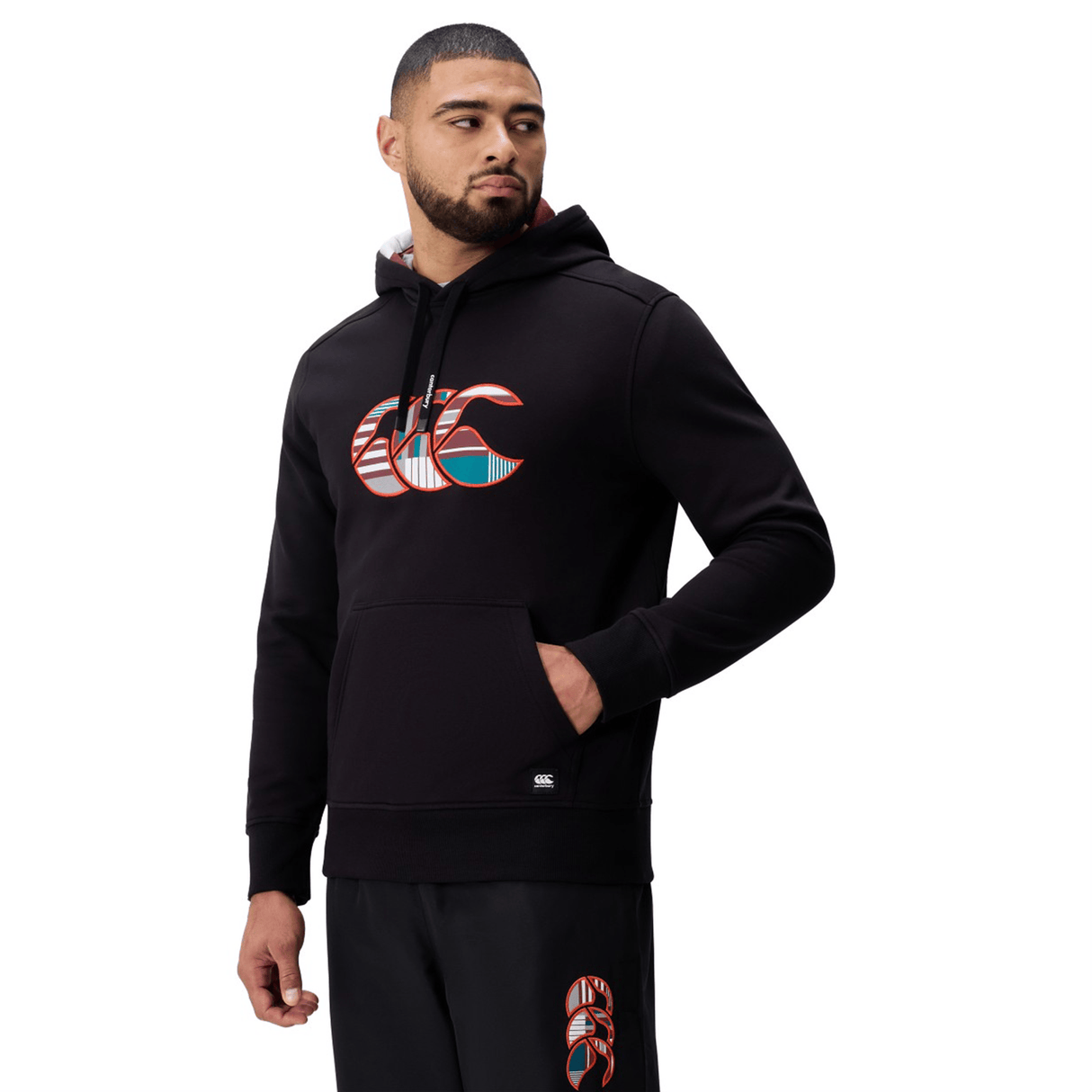 A man stands looking to his left, wearing the Canterbury Uglies Logo Hoodie in black by Canterbury and matching black pants.