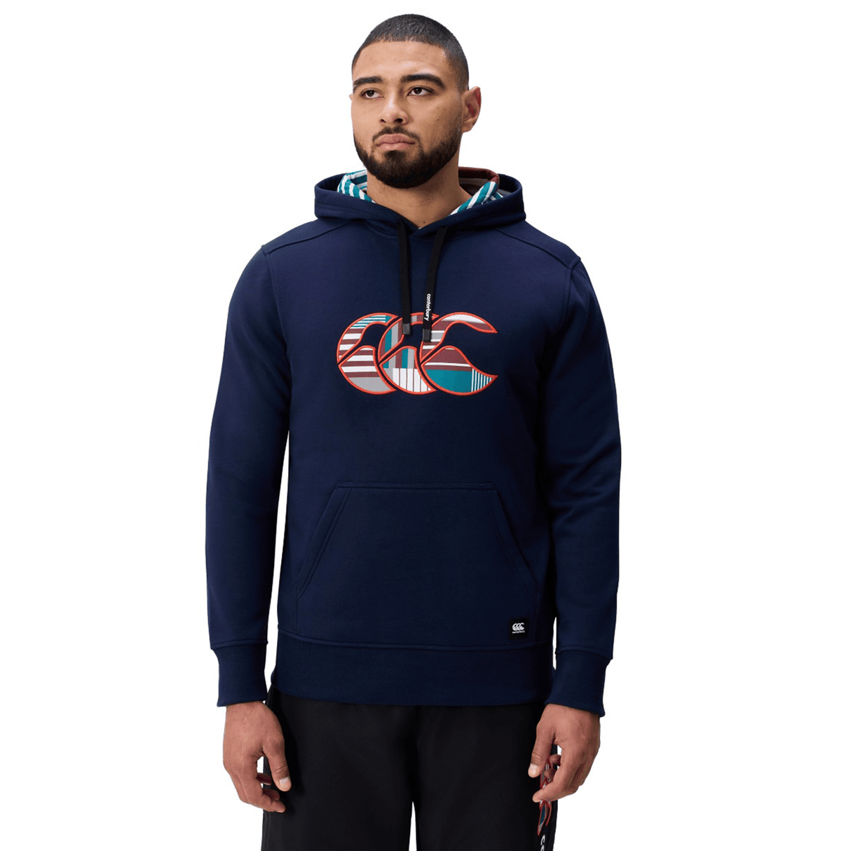 A man wearing a Canterbury Uglies Logo Hoodie in navy blue, featuring the patterned Canterbury logo on the chest, paired with black pants and standing against a white background.