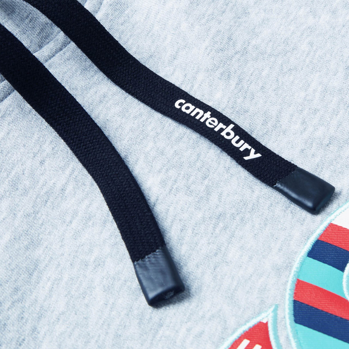 Close-up of the Canterbury Uglies Logo Hoodie drawstrings, featuring "canterbury" text on one lace, resting on light gray fabric with a glimpse of the colorful embroidered logo—a signature detail reflecting Canterbury’s bold rugby style.
