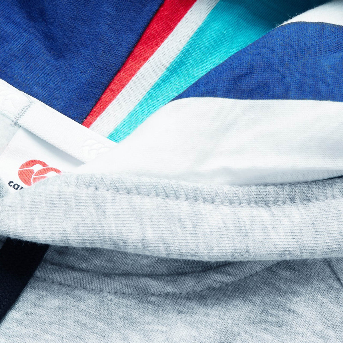 A close-up view of colorful layered fabric—featuring blue, teal, red, white, and gray segments reminiscent of the vibrant designs found on a Canterbury Uglies Logo Hoodie by Canterbury—with a partially visible clothing tag displaying printed text and the distinctive red Uglies Logo.