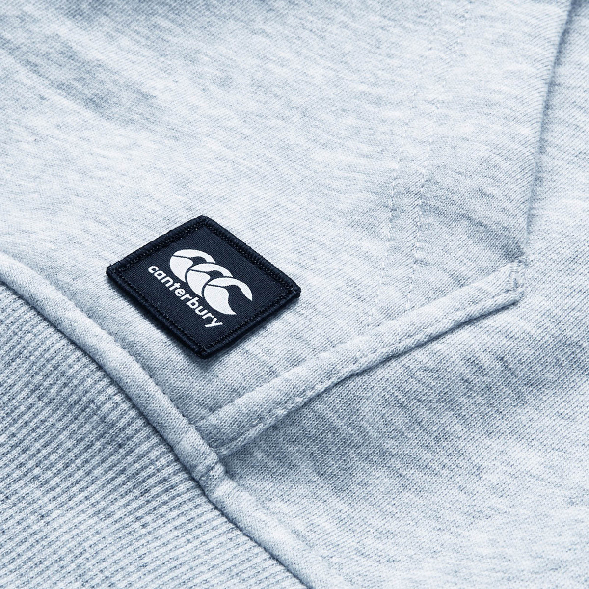 Close-up of a light gray Canterbury Uglies Logo Hoodie displaying a stitched Canterbury logo patch with three white overlapping C's on a black square background.