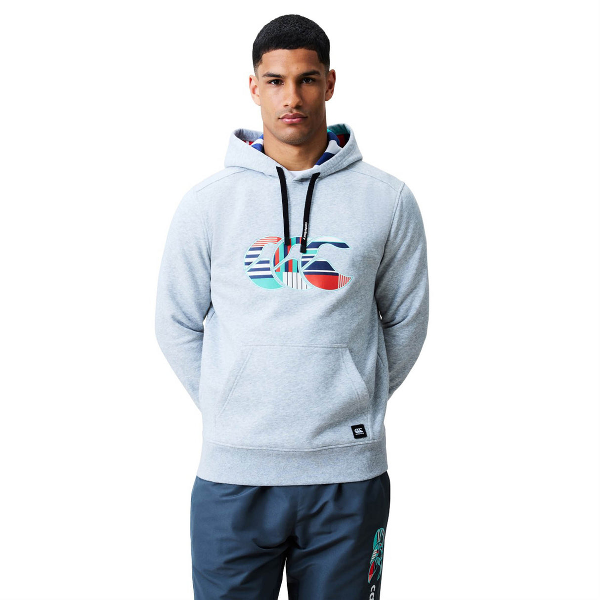 A man stands facing forward against a plain white background, wearing the Canterbury Uglies Logo Hoodie in light gray by Canterbury, featuring a colorful logo graphic on the front, paired with matching gray athletic pants.