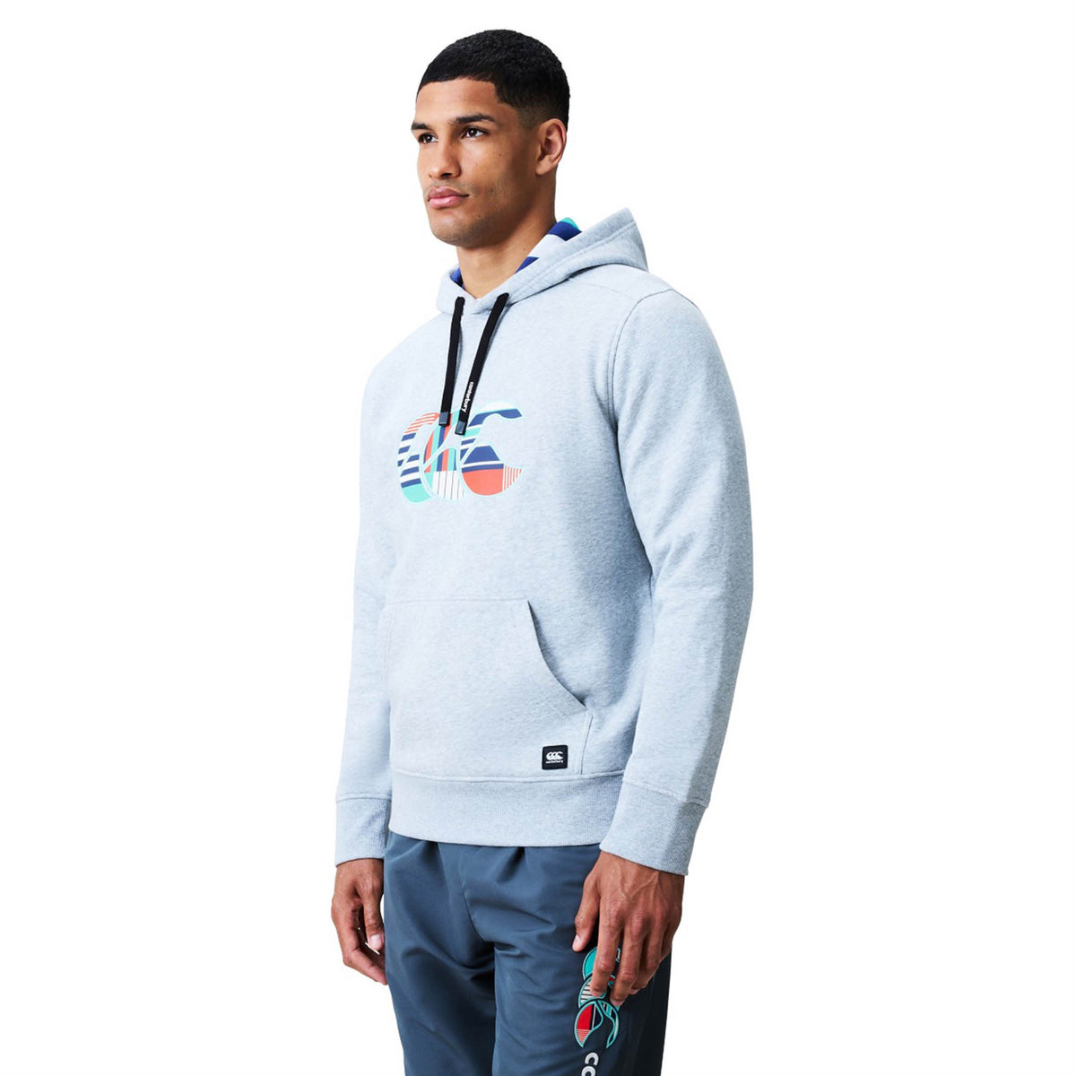 A man wearing the Canterbury Uglies Logo Hoodie in light gray, featuring a colorful graphic logo, along with matching gray sweatpants, stands facing slightly left against a plain white background.