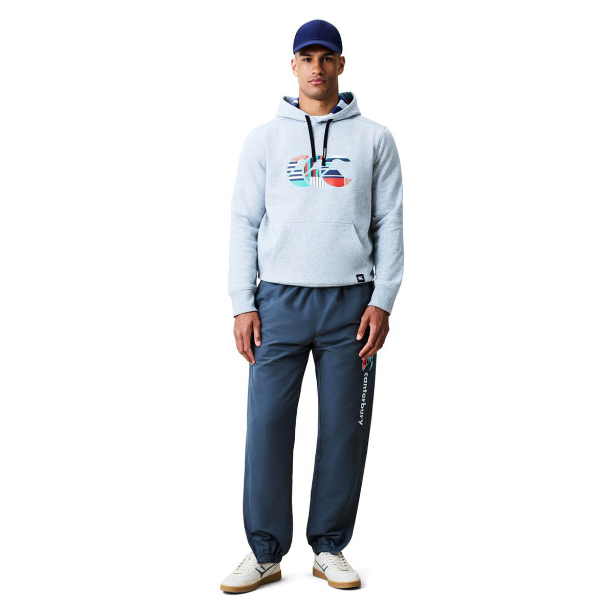 A man stands against a plain white background wearing a blue baseball cap, the Canterbury Uglies Logo Hoodie in gray with a colorful logo design by Canterbury, blue track pants, and white sneakers.