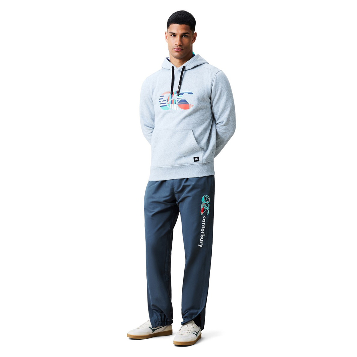 A man stands facing forward wearing a light gray hoodie with a colorful graphic, Canterbury Uglies 32" Stadium Pants by Canterbury designed for athletic performance, and light-colored sneakers.