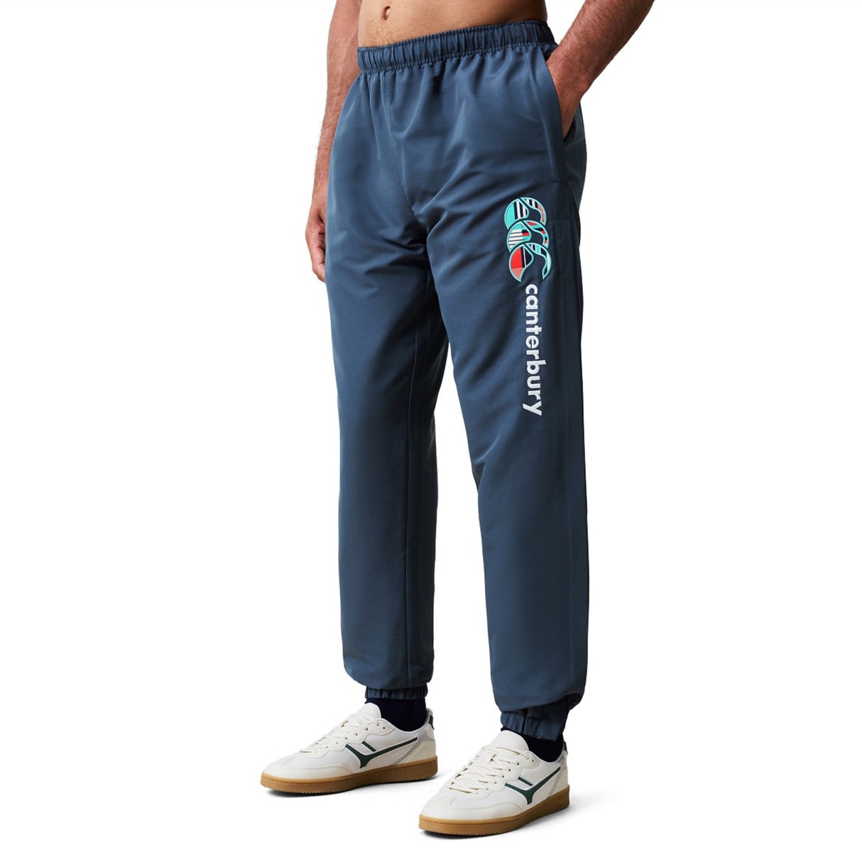 A person wears blue Canterbury Uglies 32" Stadium Pants, featuring a logo on the left leg, paired with white sneakers and a bare torso. These Canterbury pants are designed for comfort and athletic performance.