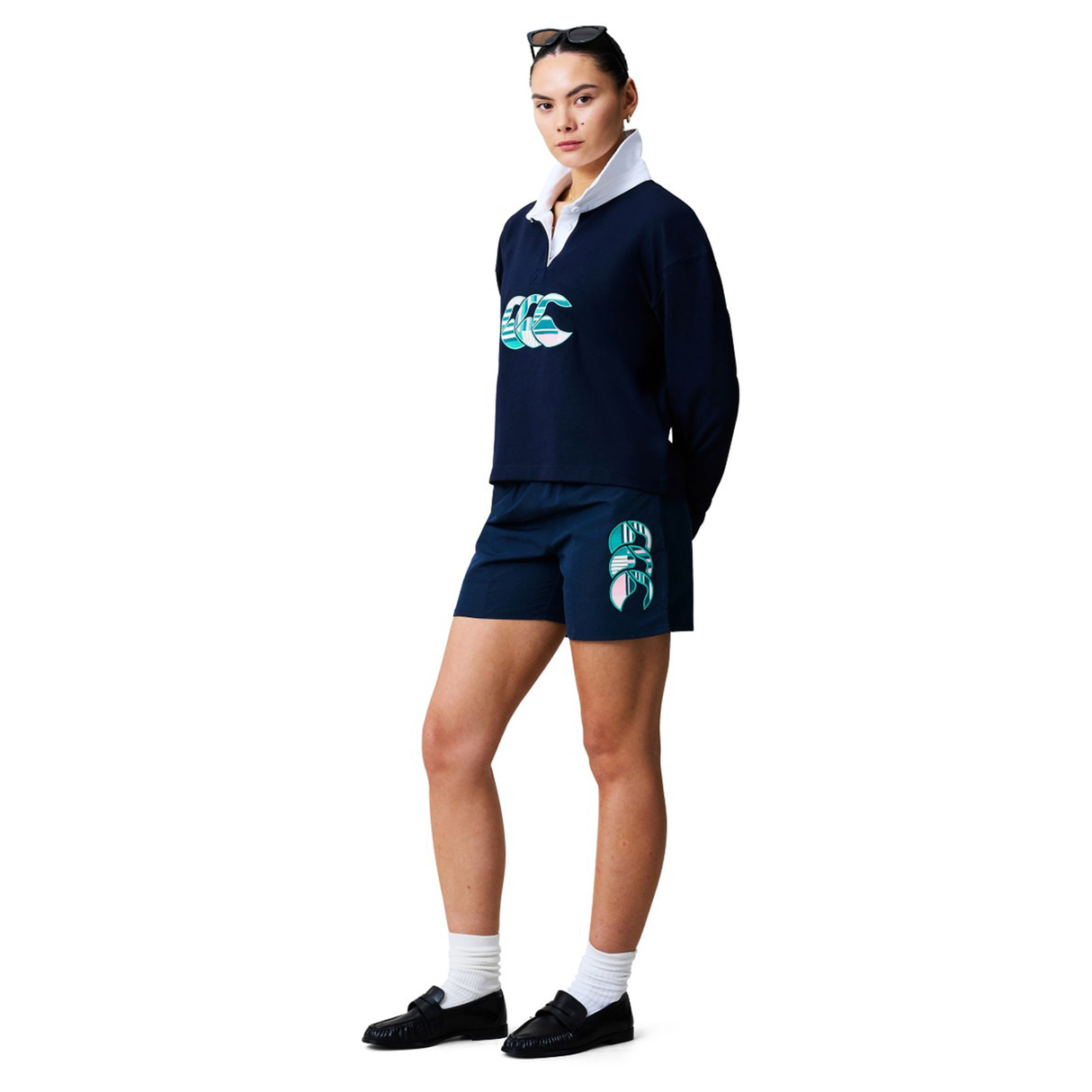 A person stands wearing a navy blue long-sleeve top with a white collar, Canterbury Women's Uglies Tactic Short by Canterbury with graphic print, white socks, black shoes, and sunglasses on their head.