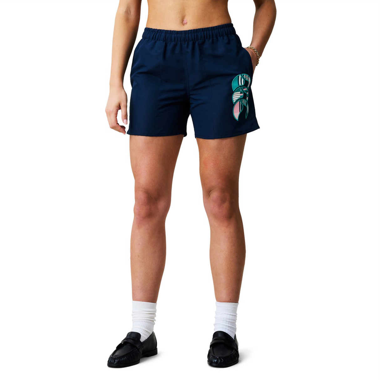 A person wears the Canterbury Women's Uglies Tactic Short in navy with a green and white leaf design on the left thigh, paired with a white top, white socks, and black shoes, standing against a plain background.