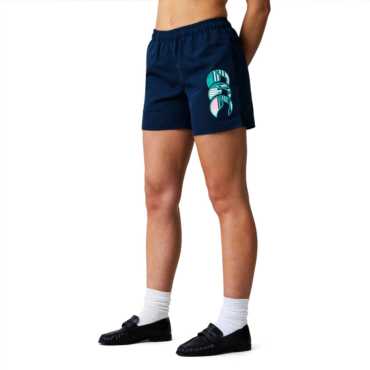 A person wears Women's Canterbury Uglies Tactic Short by Canterbury—navy blue performance shorts featuring a colorful abstract design on one leg—paired with white socks and black shoes against a plain white background.