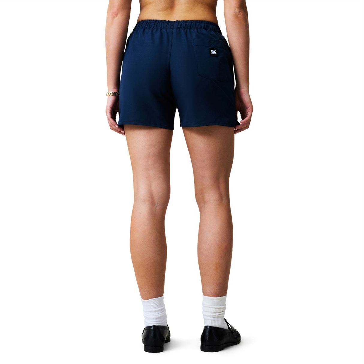 A woman stands with her back to the camera, wearing navy Canterbury Uglies Tactic Shorts, white socks, and black shoes. The shorts feature a small Canterbury logo on the right pocket.