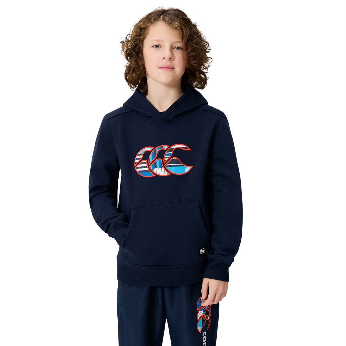 A young person with curly hair stands against a white background, hands in the kangaroo pocket of a navy Canterbury Youth Uglies Logo Hoodie and matching pants, both featuring the colorful Canterbury Uglies logo.