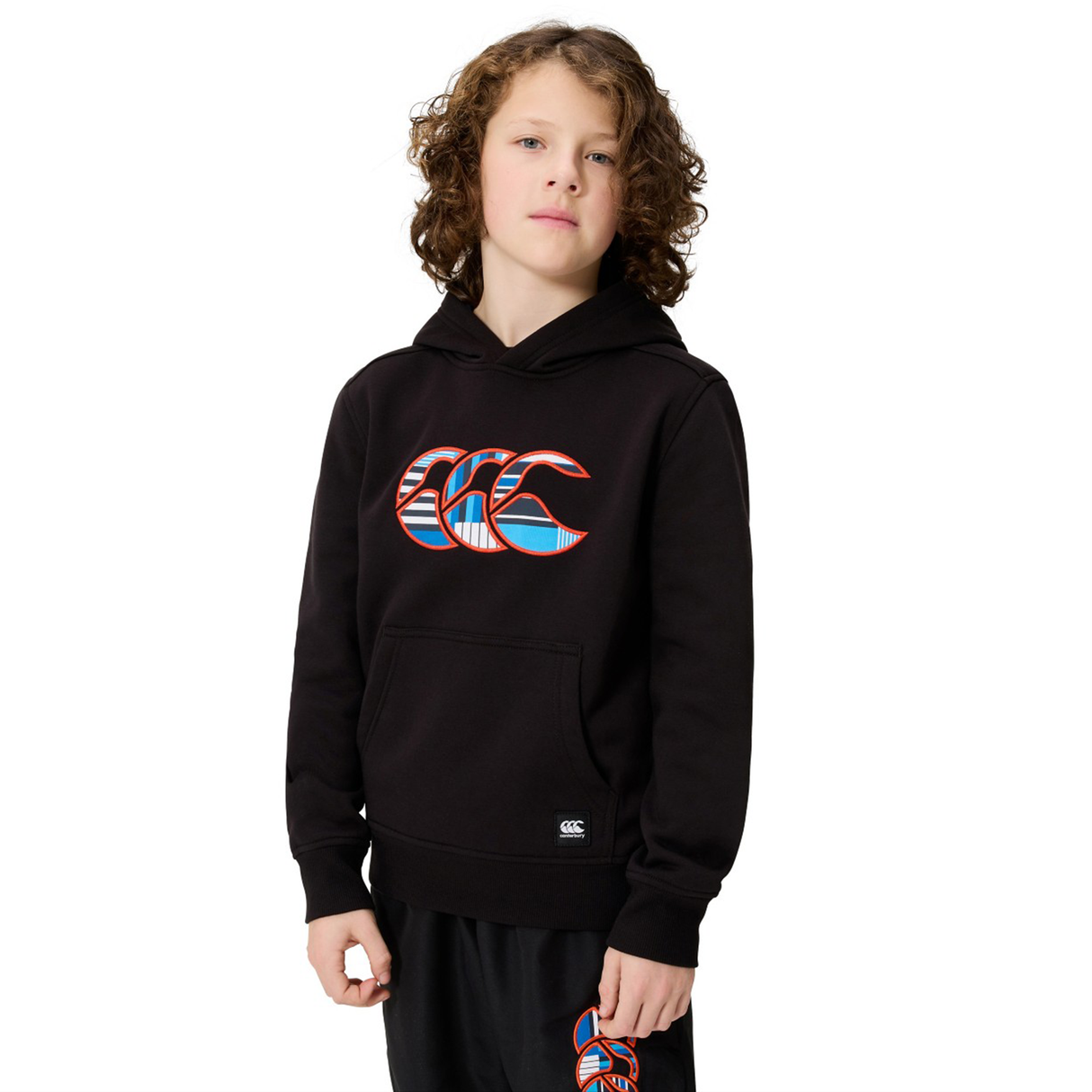 A child with curly hair stands against a plain white background wearing the Canterbury Youth Uglies Logo Hoodie in black, featuring Canterbury's iconic colorful logo, paired with matching black shorts in a similar design.
