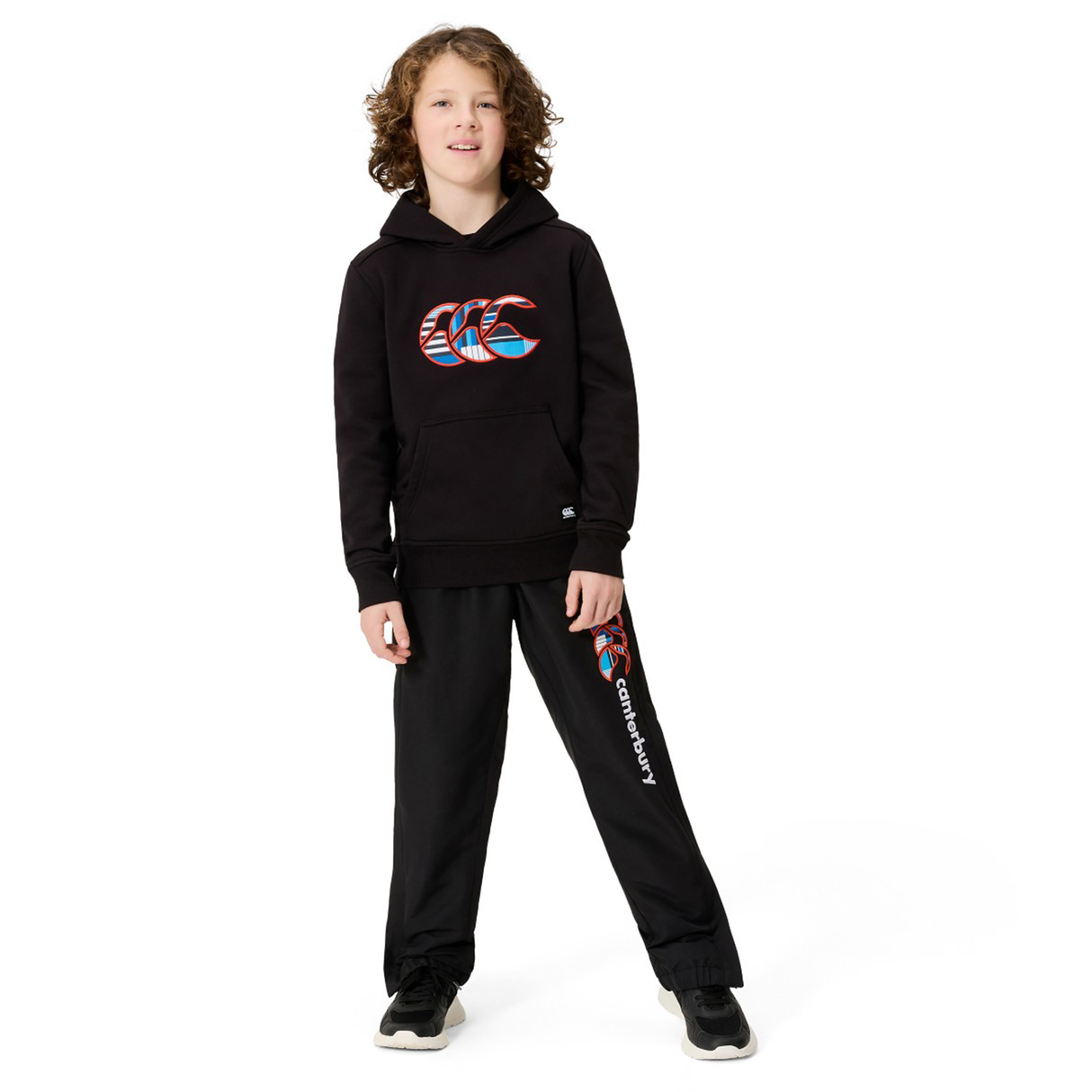 A young person with curly hair stands facing forward against a white background, wearing the Canterbury Youth Uglies Logo Hoodie in black, paired with black sweatpants featuring colorful logos, and black sneakers.