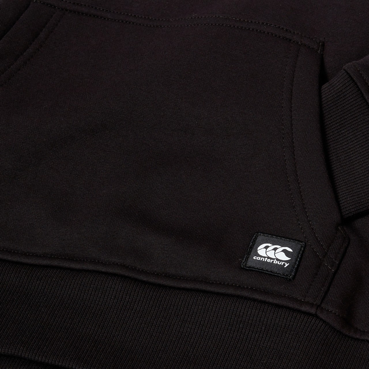 Close-up of a black Canterbury Youth Uglies Logo Hoodie showing a front pocket, ribbed hem, and a small stitched Canterbury brand label, capturing the classic rugby style.