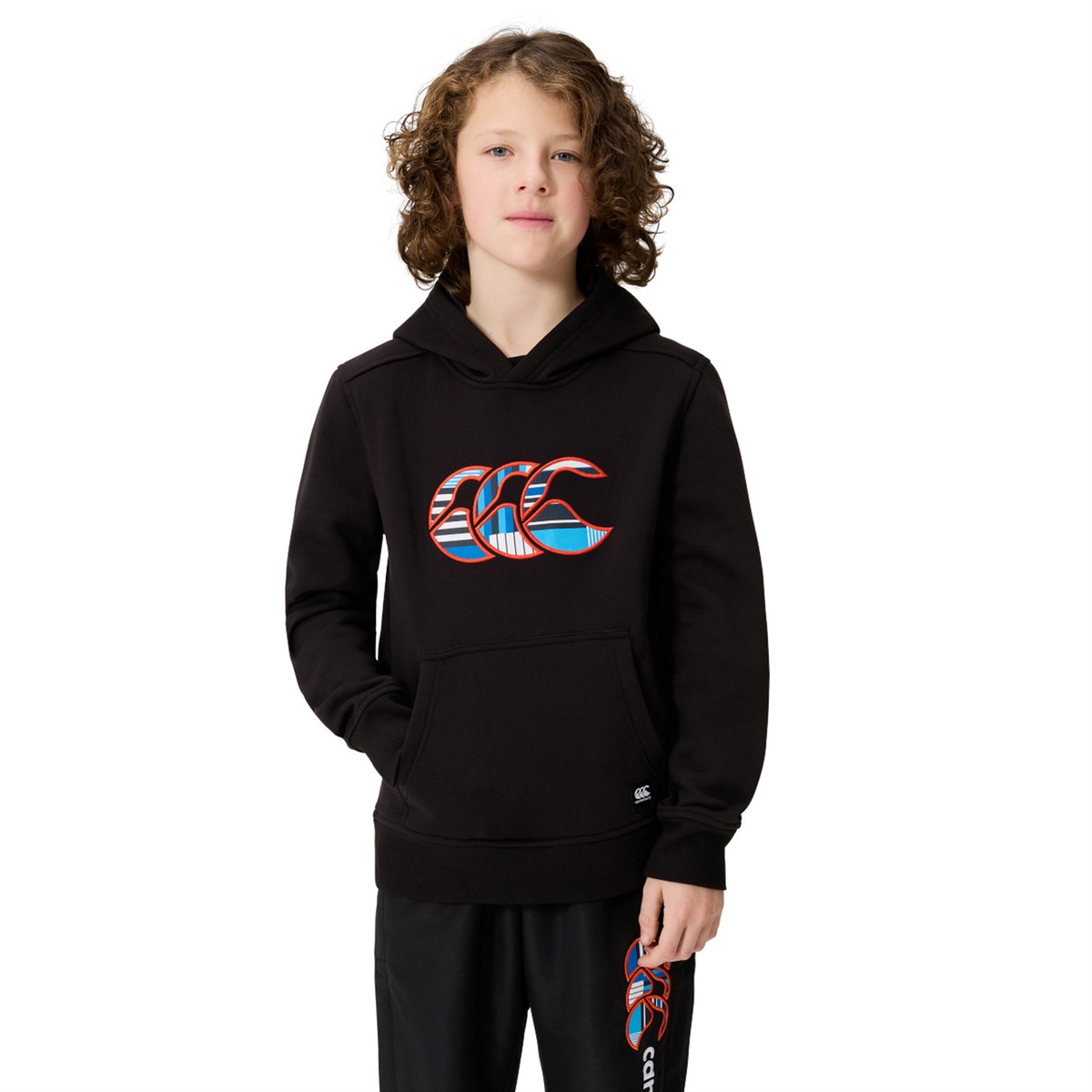 A young person with curly hair wears a black Canterbury Youth Uglies Logo Hoodie featuring a colorful wave logo that channels classic rugby style, paired with matching pants.