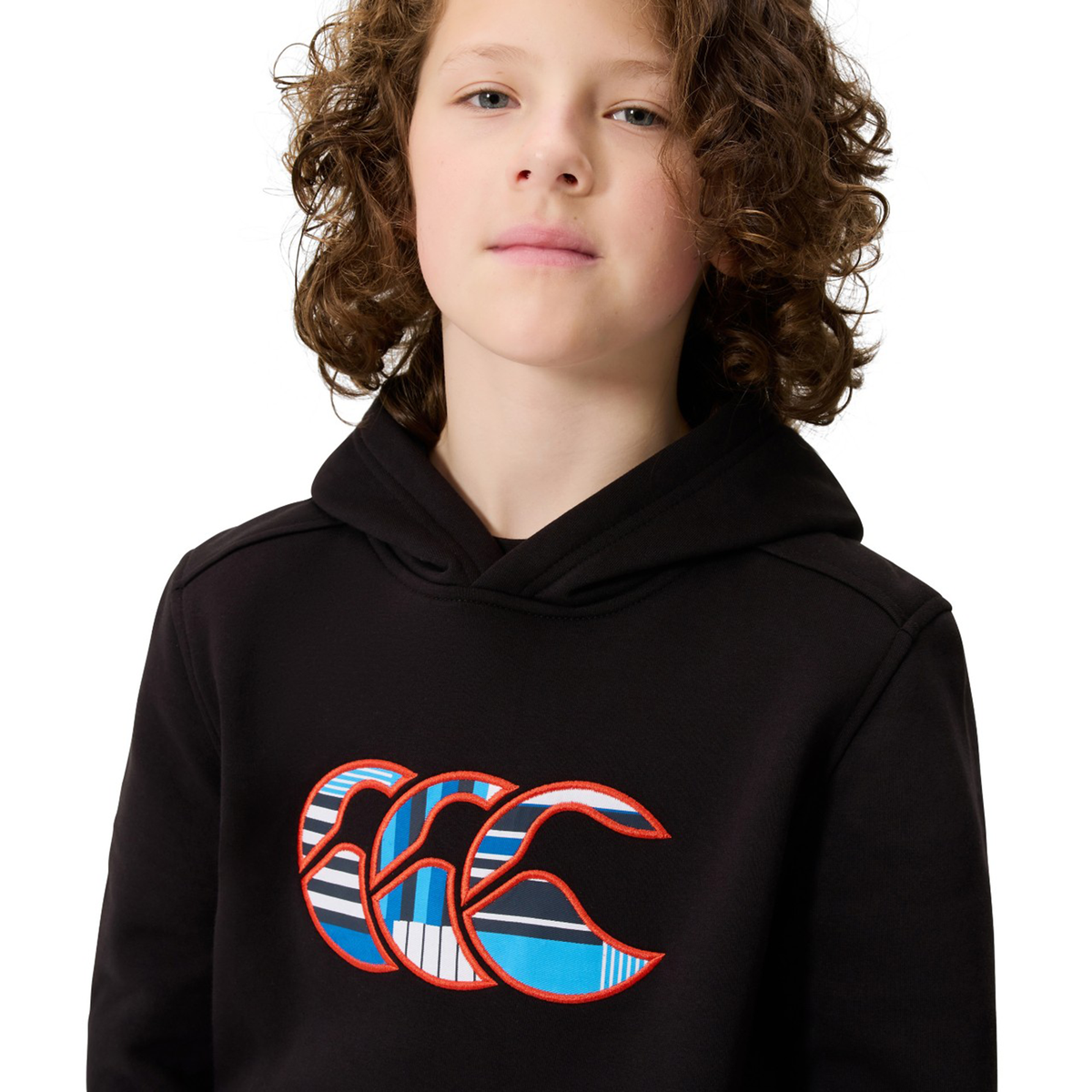 A young person with curly hair is wearing the Canterbury Youth Uglies Logo Hoodie by Canterbury, featuring a colorful "CCC" logo on the chest and shown against a white background.