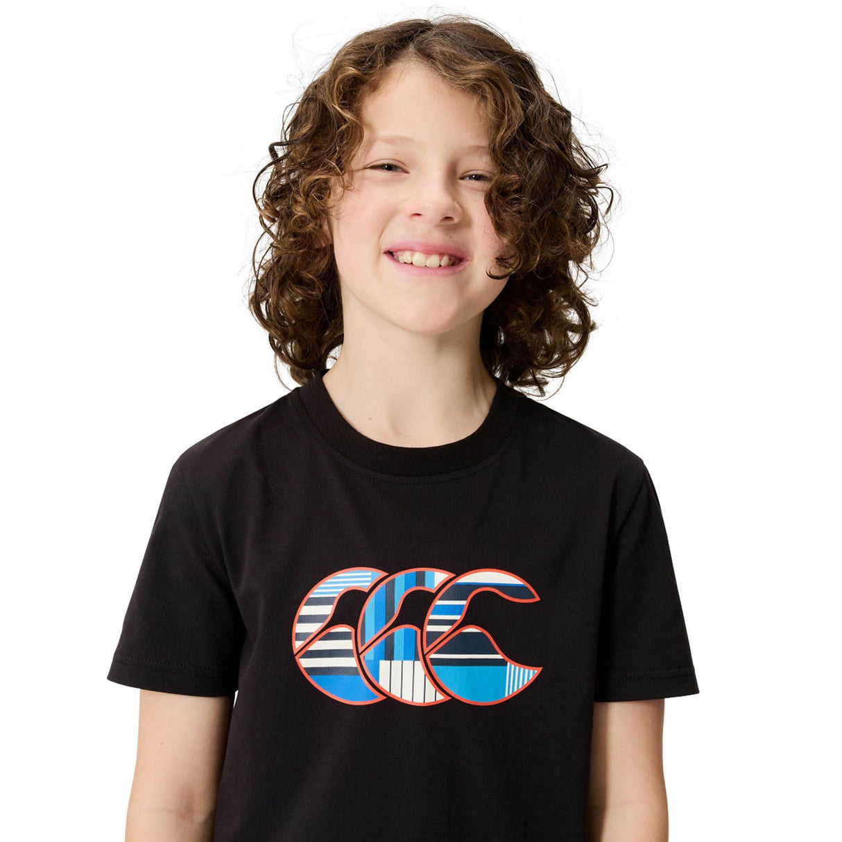 A smiling child with curly hair wears the Youth Canterbury Uglies Logo T-shirt—a comfortable cotton tee from Canterbury featuring a blue, white, and red patterned graphic design with the iconic Canterbury logo on the front—while standing against a plain white background.