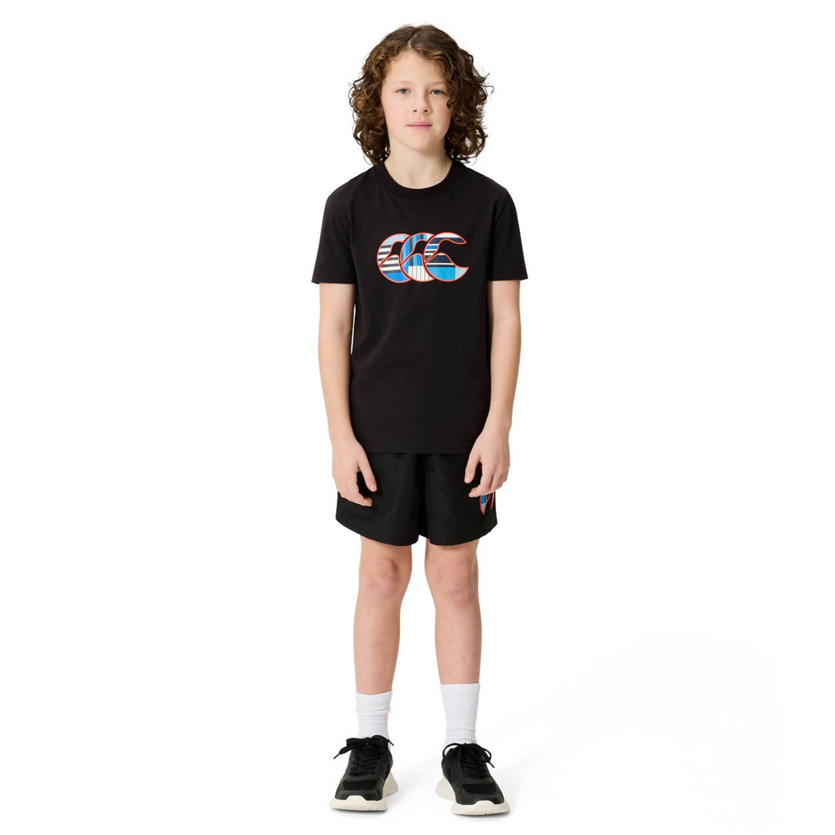 A child with curly hair stands facing forward against a white background, dressed in the Canterbury Youth Uglies Logo T-shirt in black, which showcases a vibrant Canterbury logo on the front. The t-shirt is paired with black shorts, white socks, and black sneakers. Made from comfortable cotton fabric, it offers all-day ease for active kids.