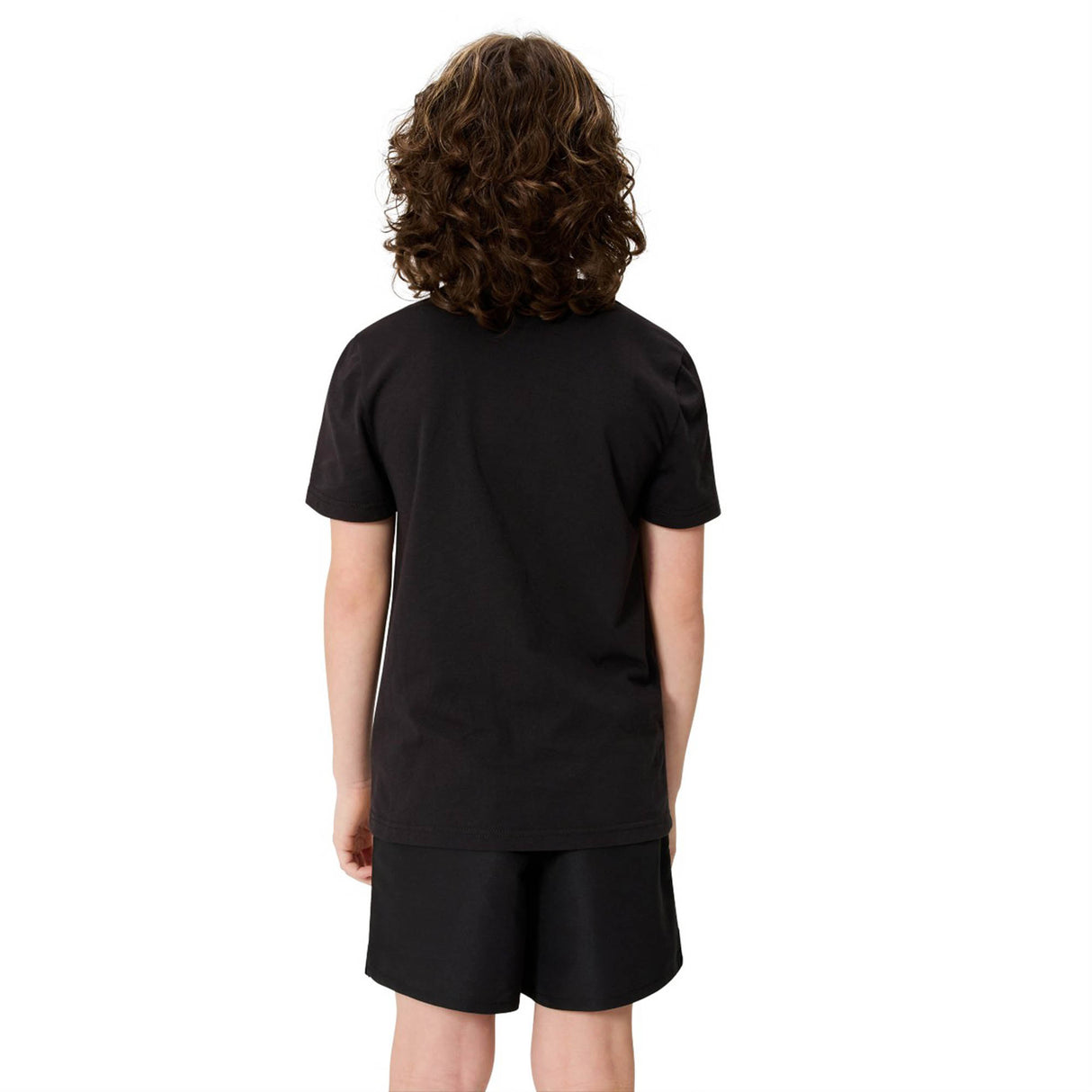 A child with curly hair stands facing away from the camera, dressed in a black Canterbury Youth Uglies Logo T-shirt made from comfortable cotton and matching black shorts, with a subtle Canterbury logo visible against a plain white background.