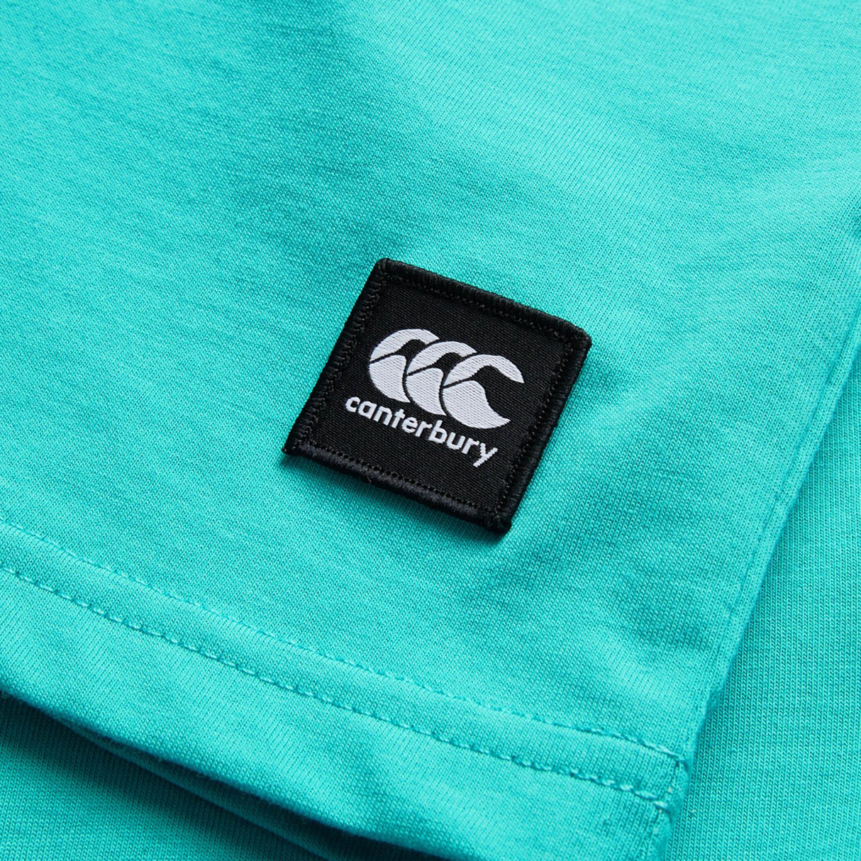 Close-up of the Youth Canterbury Uglies Logo T-shirt in turquoise, featuring a black rectangular Canterbury logo patch sewn onto the fabric.