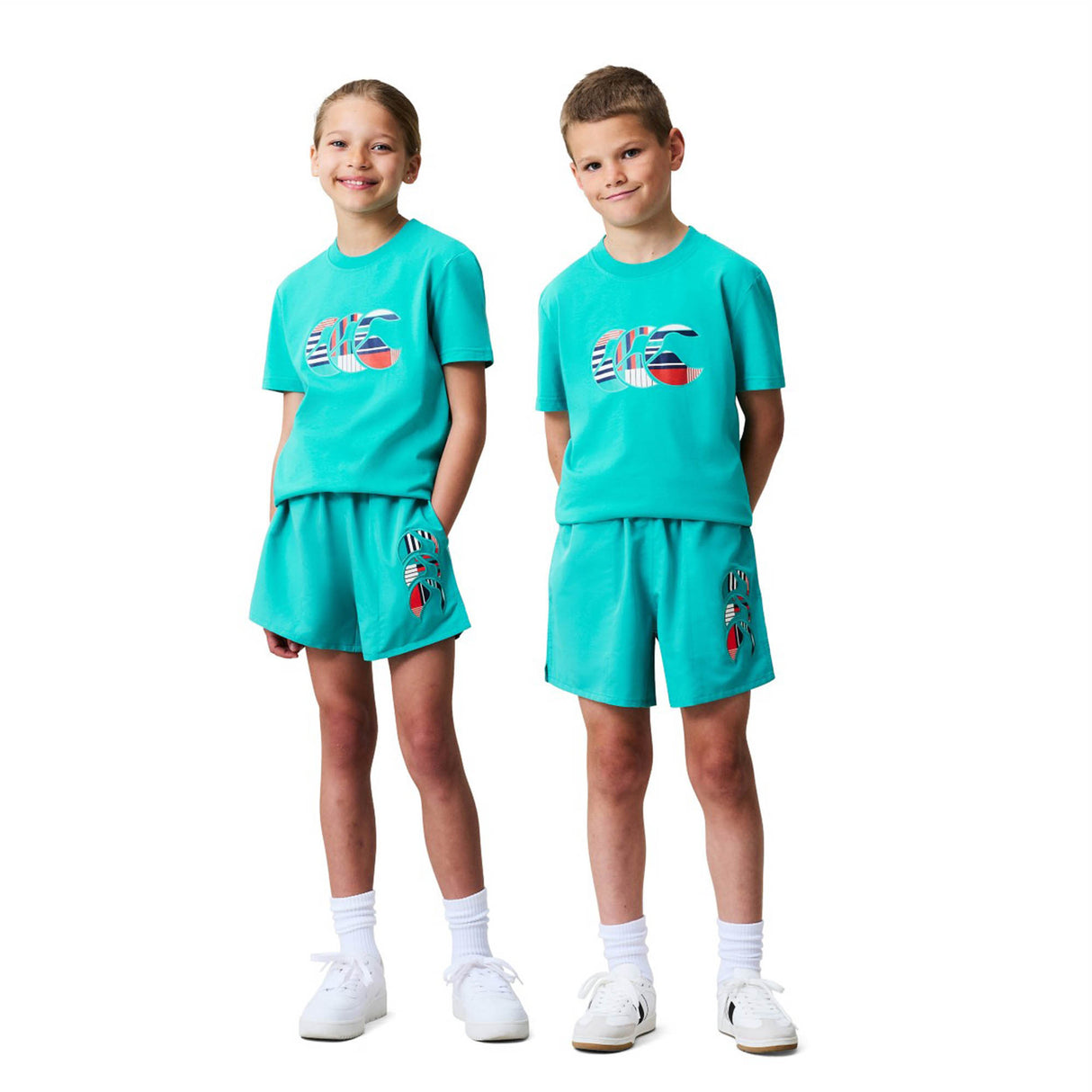 Two children stand side by side against a plain white background, both wearing matching Canterbury Youth Uglies Logo T-shirts in turquoise with colorful graphic designs, paired with shorts, white socks, and white sneakers.