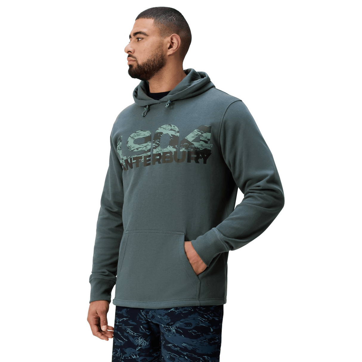 A man wearing a green Canterbury FORCE HOODIE and green patterned shorts stands facing left with one hand in his pocket.