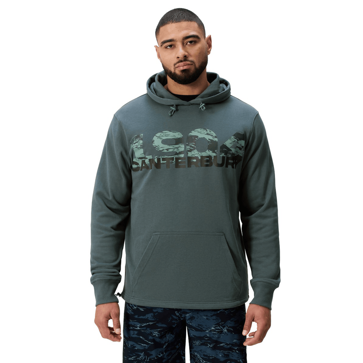 A man stands facing forward wearing a green Canterbury FORCE HOODIE with "Canterbury" and "1904" on the chest, paired with dark patterned pants.