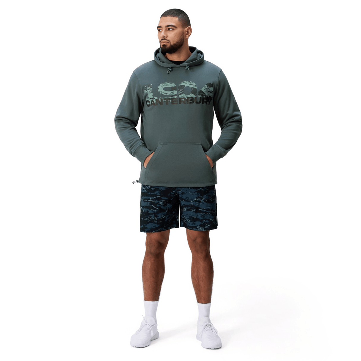 A man stands facing forward, wearing a green Canterbury FORCE HOODIE with camo print and "Canterbury" text, navy camo shorts, and white sneakers, against a plain white background.