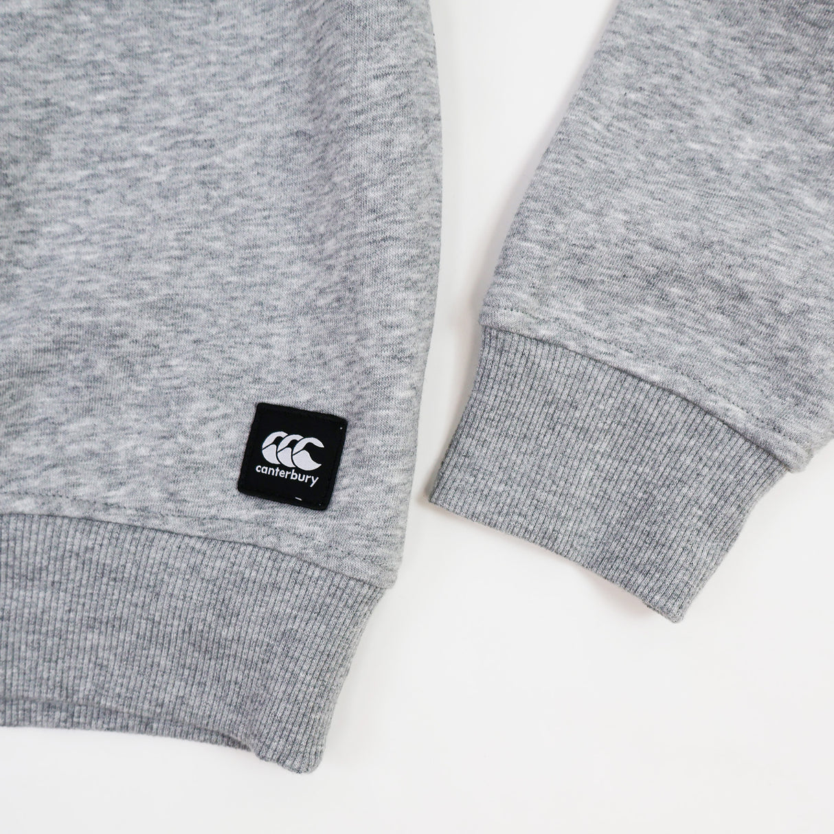 Close-up of a gray Canterbury Women's Uglies Logo Crew Sweatshirt displaying ribbed cuffs and a black Canterbury CCC logo patch near the hem on a white background.
