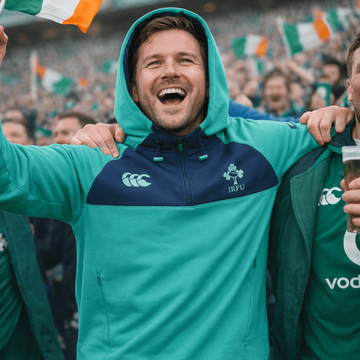 A man in a Canterbury Ireland 26/27 Summer Tour Training Hoodie smiles with arms raised as a cheering crowd with Irish flags surrounds him at a sports event.