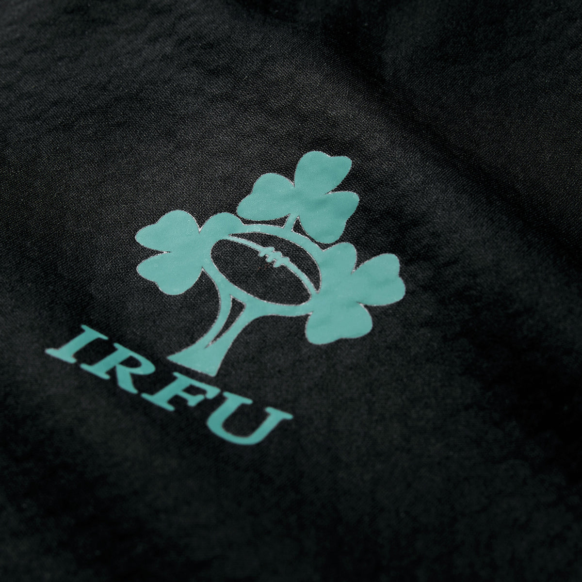 Close-up of the Ireland 26 1/4 Zip Training Top by Canterbury in black, showcasing a turquoise shamrock and rugby ball logo above “IRFU.” This Canterbury training top features innovative Thermoreg technology for enhanced comfort.