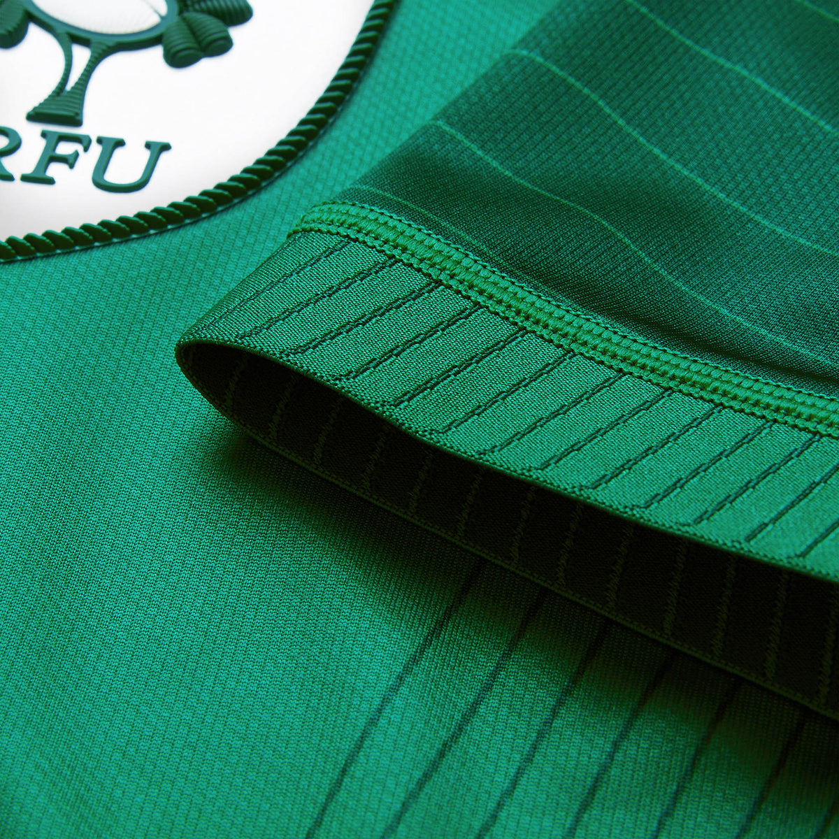 Close-up of the Youth Ireland 26 Home Pro Jersey by Canterbury in green, highlighting the edge of a sleeve with subtle stripes and part of the circular badge featuring “IRFU” lettering and a clover emblem, characteristic of an official Canterbury IRFU jersey.