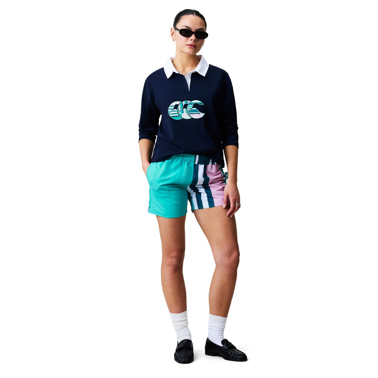 A woman wearing sunglasses, a navy collared graphic sweatshirt, Canterbury Women's Uglies Harletic Short by Canterbury in teal and pink, white socks, and black shoes stands with hands in pockets against a white background.