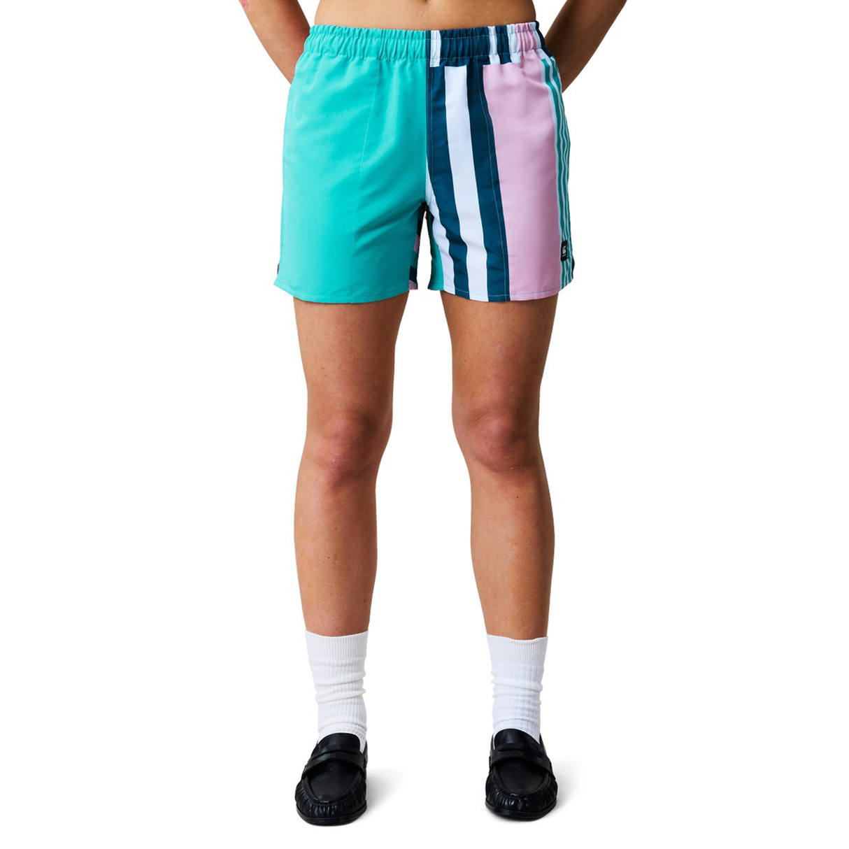 A woman wears Canterbury Women's Uglies Harletic Shorts in teal and pink with vertical stripes, paired with white socks and black shoes, standing against a white background.