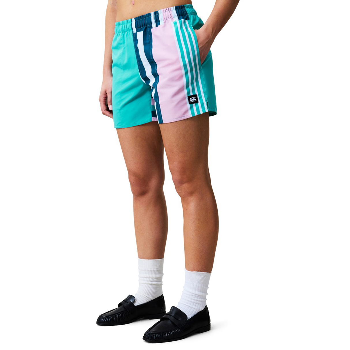 A person wears Canterbury Women's Canterbury Uglies Harletic Shorts in teal, pink, and navy stripes with white accents, paired with white socks and black loafers, standing against a white background.