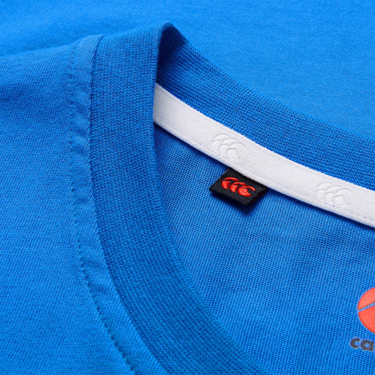 Close-up of the Canterbury Tempo+ GFX Training T-Shirt by Canterbury, a blue crew neck lightweight gym shirt featuring a visible neckline seam, a black and red tag, and a partially visible logo on the right side of the fabric.