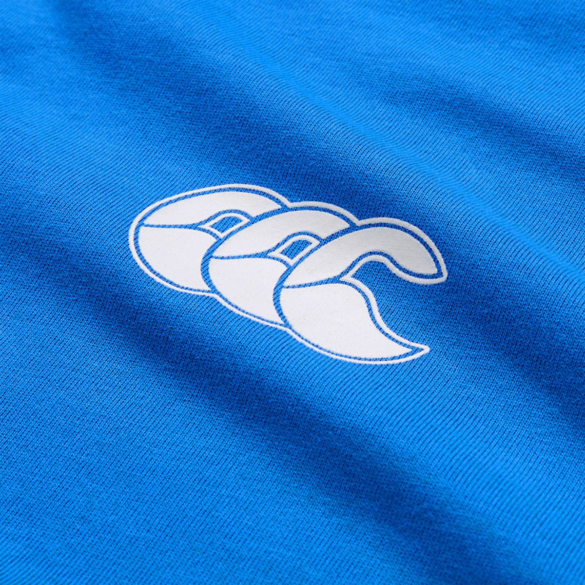 Close-up of the white Canterbury logo with three overlapping stylized “C”s printed on vibrant blue fabric, highlighting the quality and style of the lightweight Canterbury Tempo+ GFX Training T-Shirt.