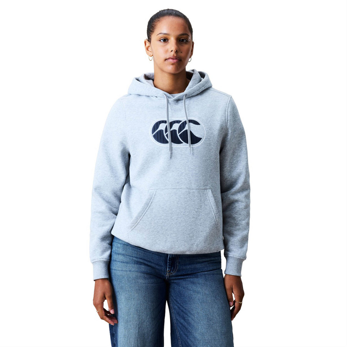 A person stands facing forward, wearing a light gray Women Rakaia 2.0 Hoodie by Canterbury featuring a dark logo, paired with blue jeans, against a plain white background.