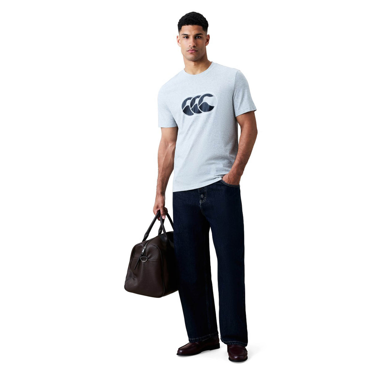 A man wearing a light gray Canterbury Rakaia 2.0 T-shirt with the Canterbury logo, paired with dark wide-leg pants and brown shoes, stands against a white background while holding a brown bag in his left hand. This stylish ensemble channels classic New Zealand rugby heritage for an effortlessly cool vibe.