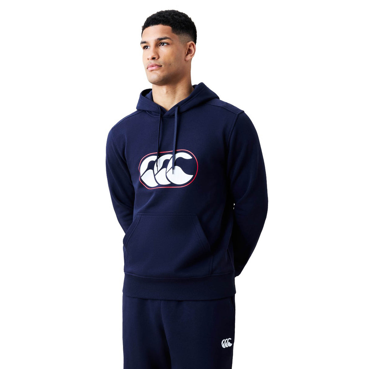 A man wearing a navy blue Canterbury Rakaia 2.0 Hoodie and matching pants, featuring a white Canterbury logo on the chest of the hoodie and the upper left leg of the pants.