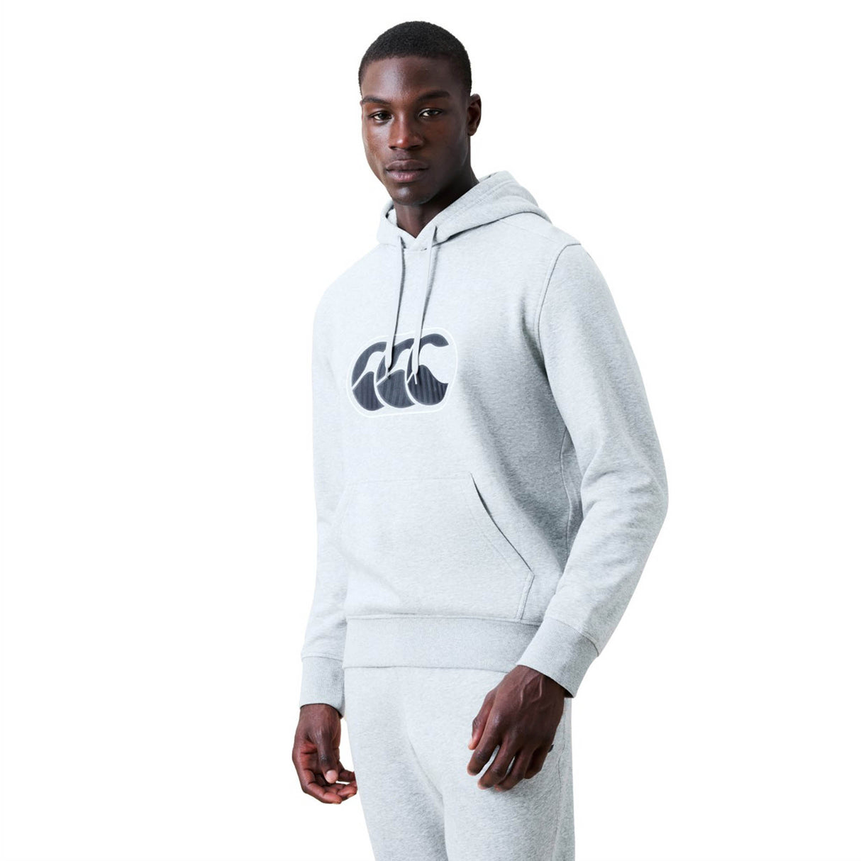 A man stands facing forward, wearing a matching set of light grey Canterbury Rakaia 2.0 Hoodie and sweatpants. The Canterbury rugby hoodie is detailed with a black logo graphic on the chest.