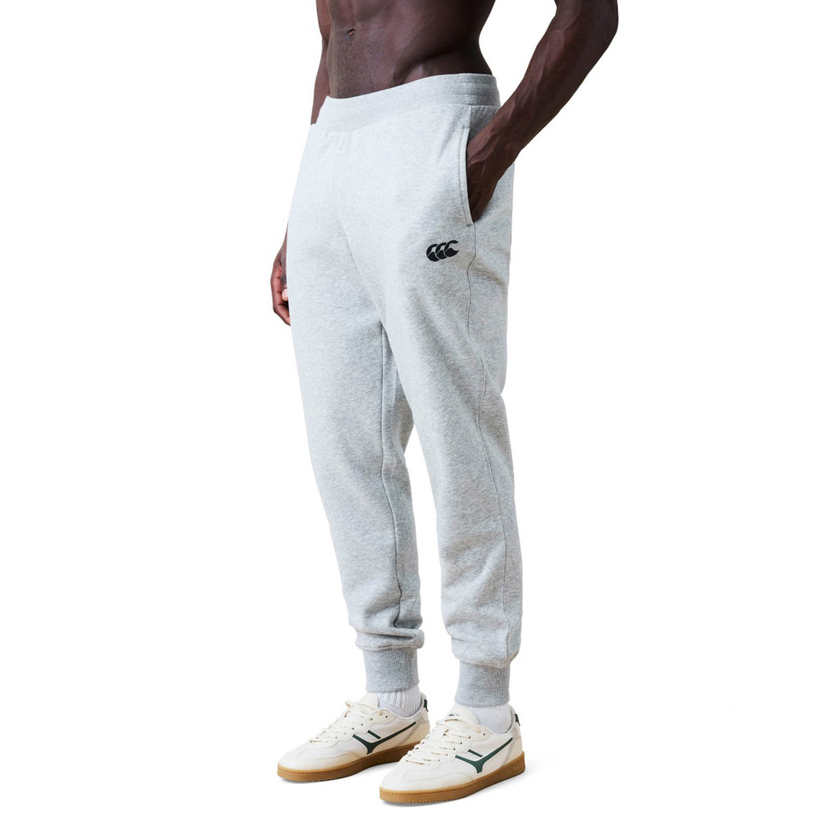 A person wearing Canterbury Rakaia 2.0 Fleece Pants in light gray with pockets and white sneakers, standing against a plain white background.