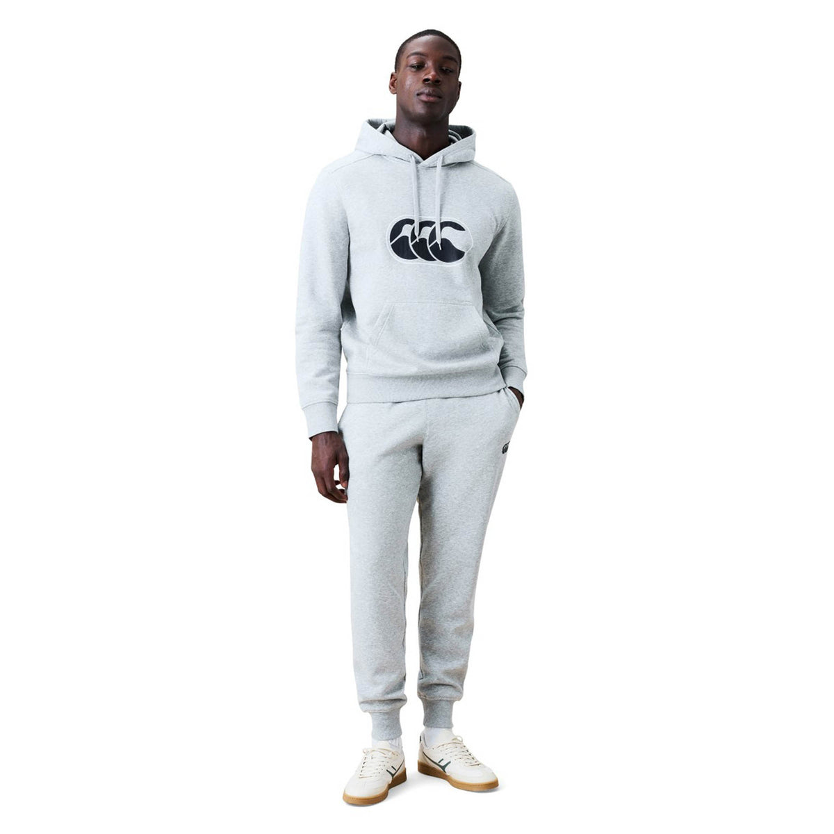 A man stands against a plain white background wearing a light gray Canterbury hoodie with a large logo, paired with Canterbury Rakaia 2.0 Fleece Pants and white sneakers.