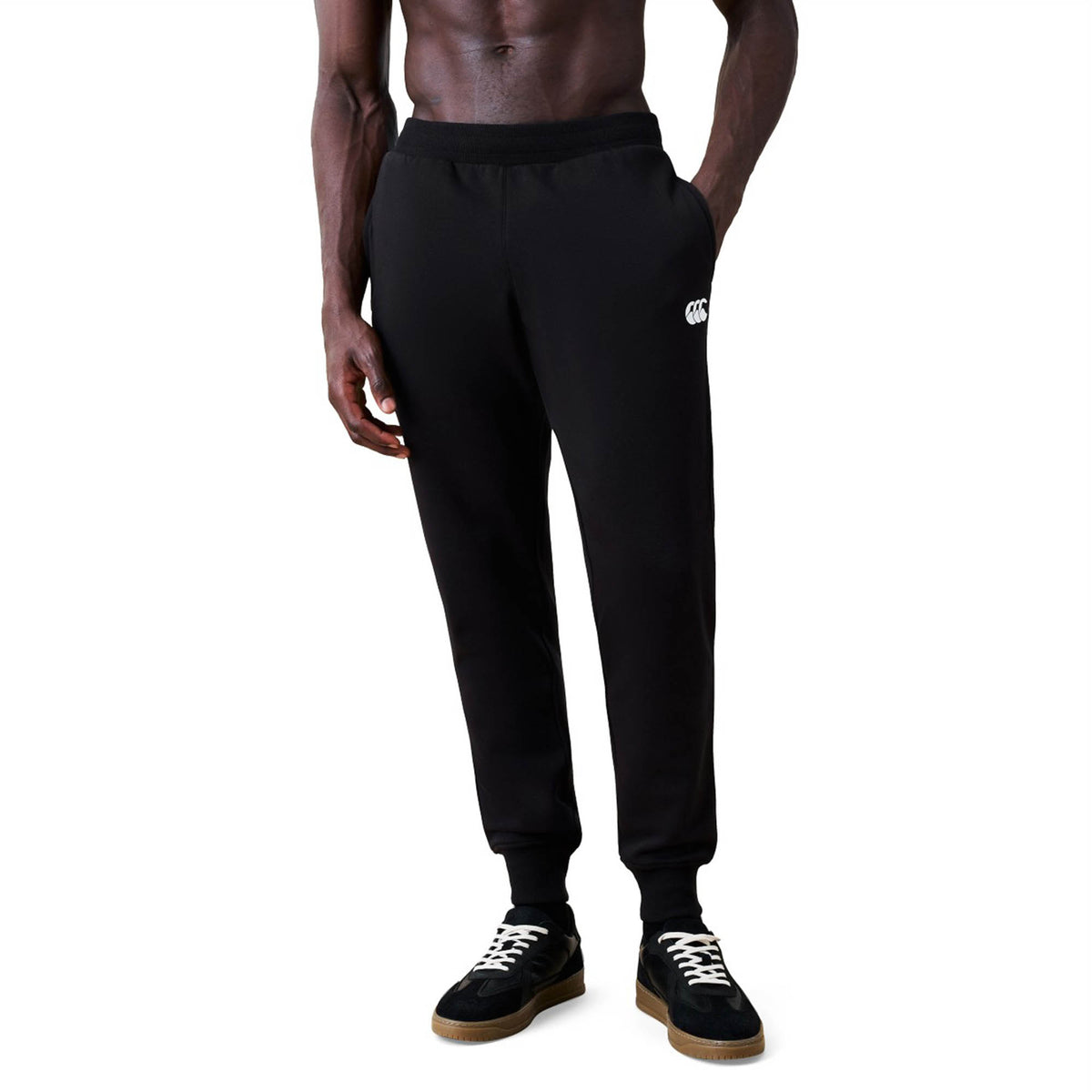 A person wearing Canterbury Rakaia 2.0 Fleece Pants in black with hands in pockets, paired with black sneakers featuring white laces and no shirt, shown from the shoulders down.