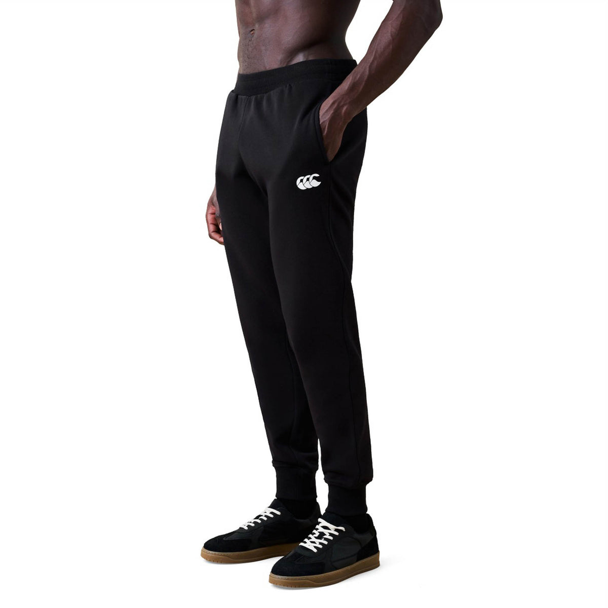 A man is wearing Canterbury Rakaia 2.0 Fleece Pant in black, featuring a white Canterbury logo on the left thigh, paired with black sneakers with white laces, while standing against a white background.