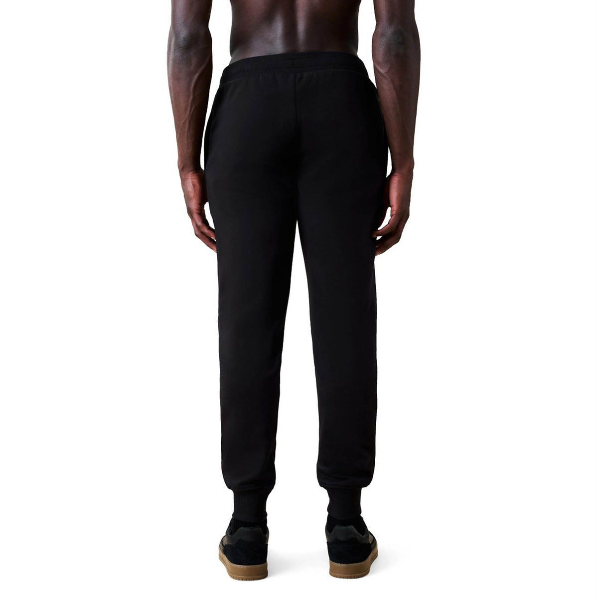 A person stands facing away from the camera, wearing Canterbury Rakaia 2.0 Fleece Pants in black and black sneakers with gum soles against a white background.