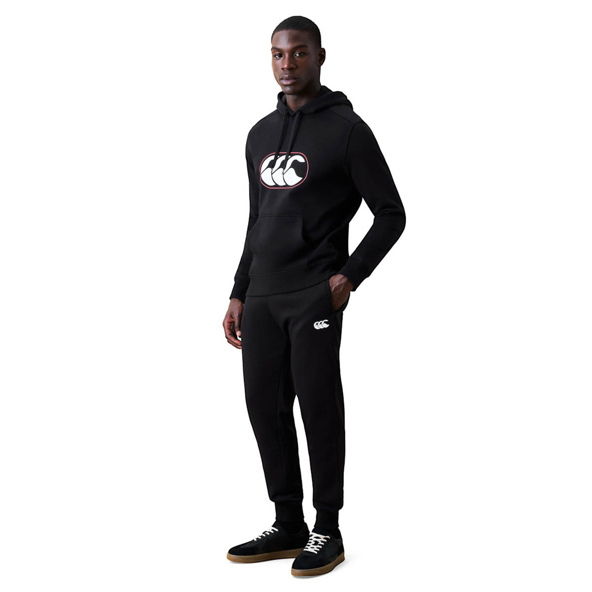 A man stands and poses in a black hoodie and Canterbury Rakaia 2.0 Fleece Pant by Canterbury, featuring white logos, paired with black sneakers on a plain white background.